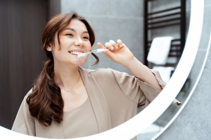 Happy,Asian,Woman,Brushing,Teeth,In,Front,Of,Bathroom,Mirror,